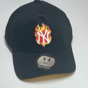 May include: Black baseball cap featuring the New York Yankees logo engulfed in flames. The logo is white and red, with yellow and orange flames. A small sticker with text is on the brim.
