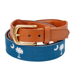 May include: A brown leather belt with a gold buckle and a blue fabric strap. The blue strap features a white tree design. The belt is a classic accessory.