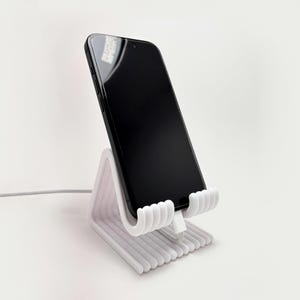 May include: A white, ribbed phone stand holding a black smartphone. The stand has a unique, modern design with a curved cradle and a base with parallel lines. A white charging cable is plugged into the phone.