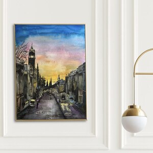 May include: A watercolor painting of a city street at sunset, with a tall building in the distance. The painting is framed and hanging on a white wall above a brown armchair with a yellow pillow. A gold floor lamp with a white globe is in the corner of the room.