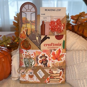 May include: A fall-themed gift box with a reading list, decorative stickers, and a cranberry snack. The box is decorated with fall leaves, a scarecrow, and a cat with the text "BooKed Up For Fall". A pumpkin-shaped jar is in the background.
