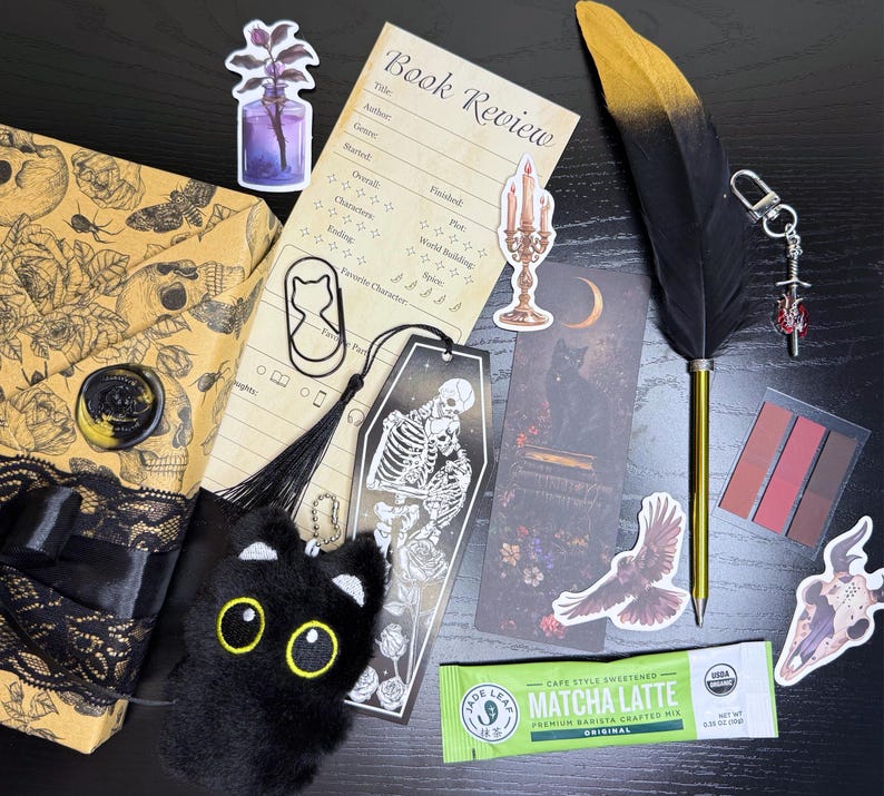 May include: A collection of gothic-themed stationery and accessories, including a black feather pen, bookmarks, stickers, a plush cat, and a wrapped gift box. The items are arranged on a dark surface, creating a moody aesthetic.