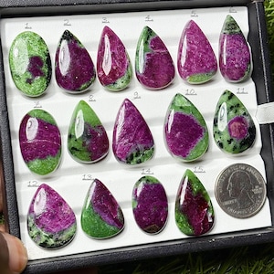 May include: A collection of fifteen polished ruby zoisite gemstones in various shapes, including oval and teardrop. The gemstones display a mix of deep magenta and green hues, with a US quarter for size comparison. The gemstones are displayed in a black tray.