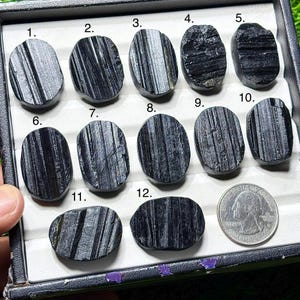 Pode incluir: Uma coleção de doze pedras de turmalina negra polidas, cada uma com estrias e texturas únicas. As pedras ovais são exibidas em uma bandeja preta, com uma moeda de 25 centavos americana para comparação de tamanho. Os detalhes da superfície variam.