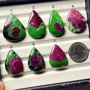 May include: Seven teardrop-shaped ruby in zoisite gemstones labeled one through seven, featuring vibrant green and deep magenta patterns. A United States quarter coin is placed next to the seventh stone for scale, providing a clear size reference.