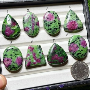 Puede incluir: Ocho piedras preciosas pulidas de rubí en zoisita con varias formas, incluyendo ovaladas, de lágrima y triangulares. Las piedras presentan una vibrante mezcla de tonos verdes y rosas, con una moneda para comparar el tamaño. Las piedras preciosas se muestran sobre una superficie blanca.