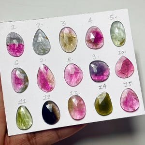 May include: A collection of fifteen faceted, pear-shaped and oval gemstones arranged on a white card, each labeled with a handwritten number from 1 to 15. The stones display a variety of colors including pink, green, yellow, and dark blue tones.