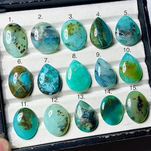 May include: A collection of fifteen polished, cabochon-cut gemstones in shades of teal, turquoise, and green, arranged in a display tray. Each stone is numbered from 1 to 15 and features unique natural inclusions, patterns, and smooth surfaces.