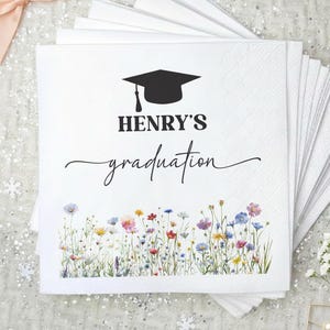 May include: White paper napkins with a graduation cap graphic and the text "HENRY'S graduation". The napkins feature a floral border with colorful wildflowers. A peach-colored bow and a small bouquet of white flowers are also visible.