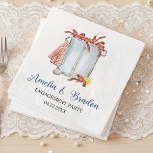 May include: White paper napkins with a watercolor illustration of a pot of crawfish, a lemon slice, and a red and white checkered towel. The text "Amelia & Bradon ENGAGEMENT PARTY 04.22.20XX" is printed in navy blue script.