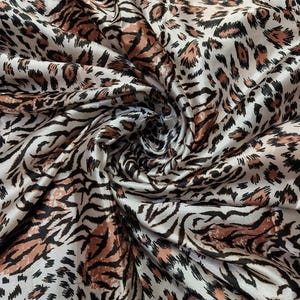 May include: A close-up of a fabric with a mixed animal print. The design combines tiger stripes in black and brown with leopard spots in black and brown, set against a cream-colored background. The fabric is gathered and folded.
