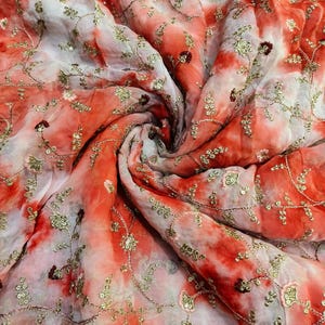 May include: A flowing fabric with a tie-dye effect in shades of red and white. The fabric is embellished with gold-colored embroidery and sequins, creating a shimmering, textured surface. The design features floral and vine patterns.