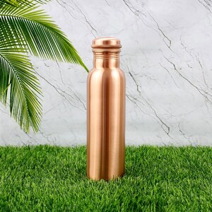 K&ouml;nnte beinhalten: Eine kupferfarbene Wasserflasche mit Schraubverschluss. Die Flasche ist zylindrisch und hat eine glatte, metallische Oberfl&auml;che. Sie steht auf gr&uuml;nem Gras, mit einem marmorierten Hintergrund.