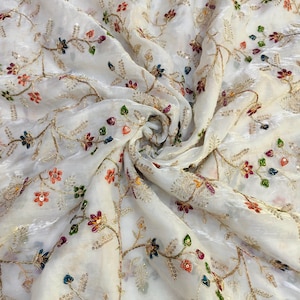 May include: White fabric with an intricate floral embroidery design. The fabric features colorful embroidered flowers in shades of red, blue, and orange, with gold-colored thread outlining the floral patterns. The fabric is slightly translucent.