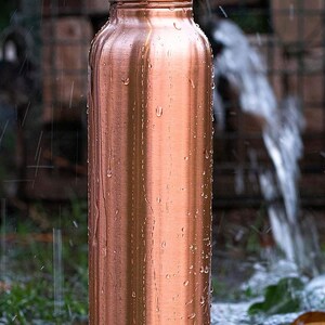 K&ouml;nnte beinhalten: Eine Kupfer-Wasserflasche mit Schraubverschluss, bedeckt mit Wassertropfen. Die Flasche hat eine zylindrische Form und steht auf gr&uuml;nem Gras. Der Hintergrund zeigt eine verschwommene Ansicht eines Wasserstrahls und Mauerwerks.