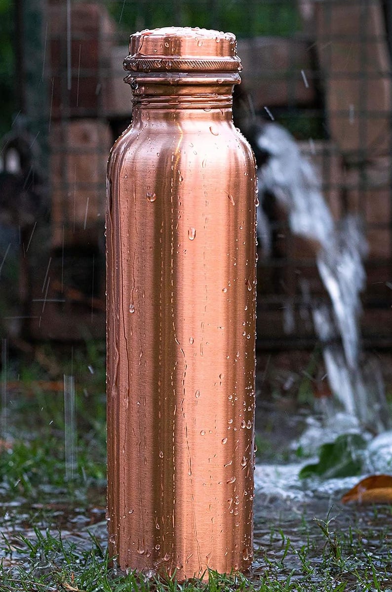 K&ouml;nnte beinhalten: Eine Kupfer-Wasserflasche mit Schraubverschluss, bedeckt mit Wassertropfen. Die Flasche hat eine zylindrische Form und steht auf gr&uuml;nem Gras. Der Hintergrund zeigt eine verschwommene Ansicht eines Wasserstrahls und Mauerwerks.