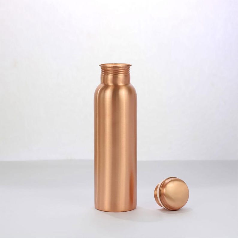 K&ouml;nnte beinhalten: Eine kupferfarbene Wasserflasche mit passendem Deckel. Die Flasche hat eine zylindrische Form mit einem Gewindehals. Der runde Deckel liegt neben der Flasche. Die Artikel sind aus gl&auml;nzendem Metall und stehen auf einer wei&szlig;en Oberfl&auml;che.