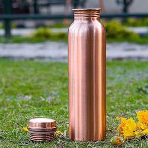 K&ouml;nnte beinhalten: Eine kupferfarbene Wasserflasche und ihr Deckel liegen auf gr&uuml;nem Gras. Die Flasche ist hoch und zylindrisch, mit einem Schraubverschluss. Der Deckel ist abgenommen und liegt neben der Flasche. Im Hintergrund sind gelbe Blumen zu sehen.