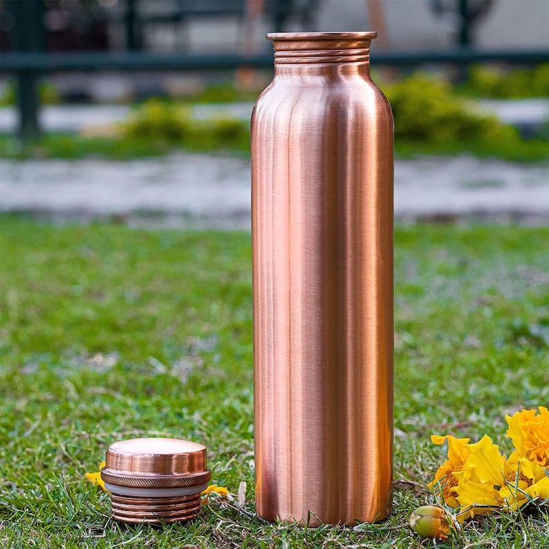 K&ouml;nnte beinhalten: Eine kupferfarbene Wasserflasche und ihr Deckel liegen auf gr&uuml;nem Gras. Die Flasche ist hoch und zylindrisch, mit einem Schraubverschluss. Der Deckel ist abgenommen und liegt neben der Flasche. Im Hintergrund sind gelbe Blumen zu sehen.