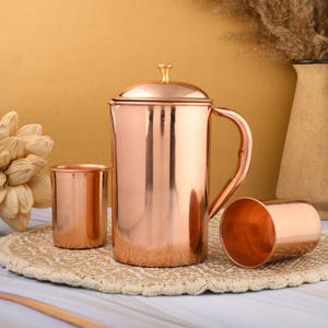 May include: A copper jug with a lid and handle, alongside two copper beakers. The set is presented on a woven mat against a warm, neutral backdrop. The jug and beakers have a polished, reflective surface, typical of copperware.