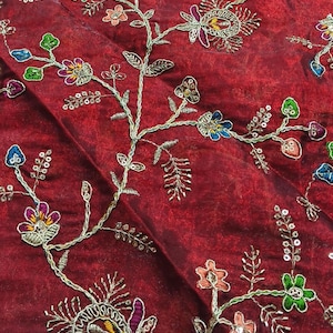 May include: Close-up of a deep red fabric with intricate floral embroidery. The embroidery features silver metallic thread and colourful accents in shades of pink, green, blue, and yellow. The design includes detailed flowers and delicate leaves.