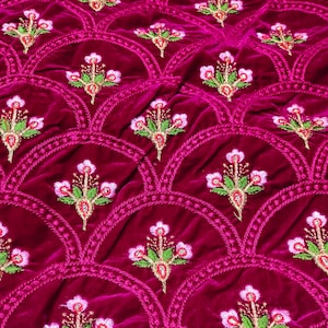 Könnte beinhalten: Nahaufnahme eines tiefmagentafarbenen Samtstoffs mit einem gestickten Blumenmuster. Das Design zeigt sich wiederholende Blumenmotive in Rosa, Grün und Gold, die in einem wellenförmigen, genähten Rand angeordnet sind. Die weiche Textur des Samts ist sichtbar.