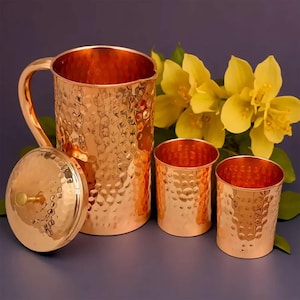 Könnte beinhalten: Ein gehämmertes Kupferkrug mit passendem Deckel und zwei Kupferbechern. Das Set wird auf einer blauen Oberfläche mit gelben Blumen im Hintergrund präsentiert. Das Kupfer hat eine glänzende, reflektierende Oberfläche.