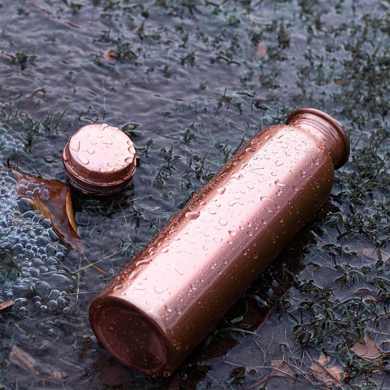 K&ouml;nnte beinhalten: Eine Kupfer-Wasserflasche mit passendem Deckel, die auf einer nassen Oberfl&auml;che liegt. Die Flasche ist zylindrisch mit leicht strukturierter Oberfl&auml;che und mit Wassertropfen bedeckt. Der Deckel ist abgenommen und liegt daneben.