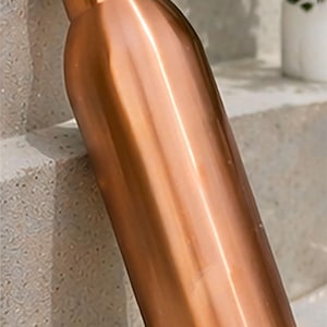 May include: A copper-colored water bottle with a rounded body and a screw-on lid. The bottle is leaning against a concrete surface. The bottle is approximately 10 inches tall.