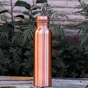 K&ouml;nnte beinhalten: Eine kupferfarbene Wasserflasche mit Schraubverschluss. Die Flasche hat eine zylindrische Form und eine glatte, polierte Oberfl&auml;che. Der Hintergrund zeigt gr&uuml;nes Laub und eine Steinoberfl&auml;che.