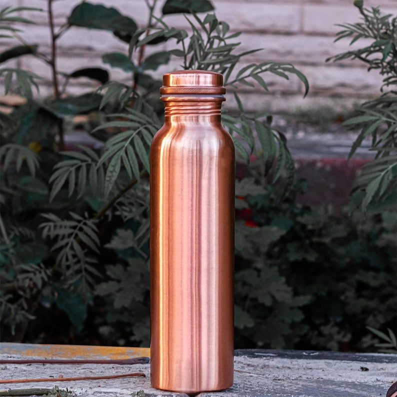 K&ouml;nnte beinhalten: Eine kupferfarbene Wasserflasche mit Schraubverschluss. Die Flasche hat eine zylindrische Form und eine glatte, polierte Oberfl&auml;che. Der Hintergrund zeigt gr&uuml;nes Laub und eine Steinoberfl&auml;che.