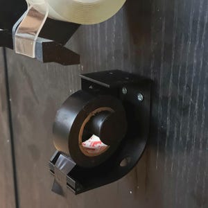 May include: Black tape dispenser with a roll of black electrical tape and a roll of clear tape. The dispenser is mounted on a dark wooden surface. The tape is ready to be used.