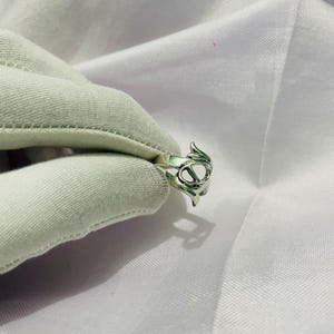 以下が含まれることがあります： ユニークなデザインのシルバーリング。尖ったエッジを持つ、様式化されたオープンな形状が特徴です。リングは白い布の背景に表示され、その複雑なディテールと職人技を際立たせています。