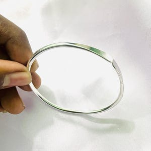 Może przedstawiać: Srebrna bransoletka typu obręcz o prostym, eleganckim designie. Bransoletka jest okrągła z płaskim, prostokątnym fragmentem. Metal ma błyszczącą, odbijającą światło powierzchnię. Bransoletka jest trzymana przez dłoń na białym tle.