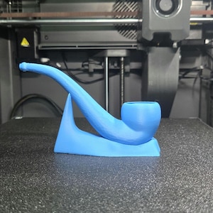 May include: A light blue, 3D-printed tobacco pipe. The pipe features a curved stem and a bowl, resting on a matching base. The object is made of plastic and sits on a dark surface.