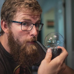 May include: A clear glass pipe with a spherical bowl and a black stem. The pipe is held by a person with a beard and glasses. The bowl of the pipe is filled with small, reflective objects.