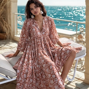 May include: A flowing, rust-colored dress with a white floral pattern. The dress features a V-neck and three-quarter sleeves. The model is wearing shell earrings and a gold necklace. The background shows a view of the ocean.