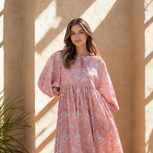 May include: A long, flowing dress with a floral pattern in shades of coral and gray. The dress features long, puffy sleeves and a gathered waist. The model is standing in front of a textured wall, with sunlight casting shadows.