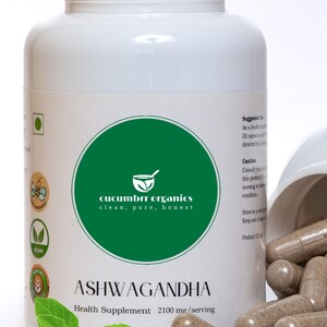 May include: A white bottle of Ashwagandha health supplement with a green label featuring the text "cucumbrr organics". Brown capsules are spilled from an open container. A sprig of green leaves and some roots are in the foreground.