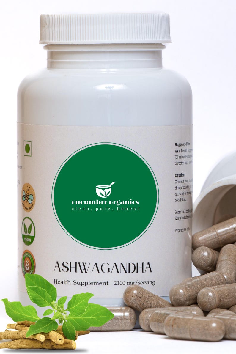 May include: A white bottle of Ashwagandha health supplement with a green label featuring the text "cucumbrr organics". Brown capsules are spilled from an open container. A sprig of green leaves and some roots are in the foreground.