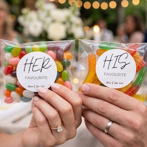 May include: Two clear plastic bags filled with sweets. The left bag contains multi-coloured jelly beans with a white circular label reading HER FAVOURITE Alexa & John 2026. The right bag contains gummy worms with a white label reading HIS FAVOURITE Alexa & John 2026.