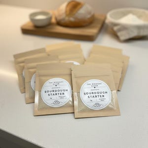 May include: A collection of small, tan sourdough starter pouches with clear windows and circular labels reading "SOURDOUGH STARTER" and "THE MIDNIGHT BAKER". A loaf of bread, a small bowl of salt, and a basket of flour are in the background, on a white surface.