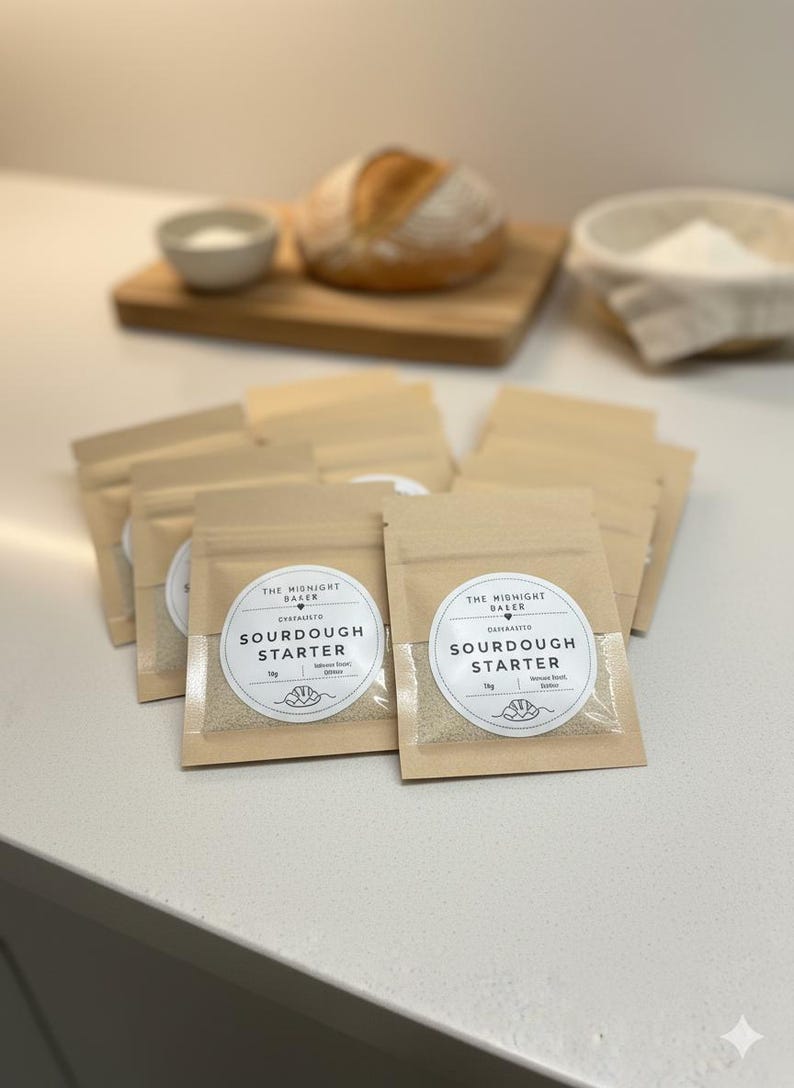 May include: Several small, tan pouches of sourdough starter are arranged on a white surface. Each pouch has a clear window and a circular label that reads "SOURDOUGH STARTER" and "THE MIDNIGHT BAKER". A loaf of bread, a bowl of salt, and a basket of flour are in the background.
