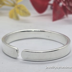 Könnte beinhalten: Ein silberfarbener, verstellbarer Ring mit flachem Band und offenem Design. Der Ring wird auf hellem Hintergrund mit verschwommenen Blumen gezeigt. Der Text "jewellrybyhandmade" ist sichtbar.