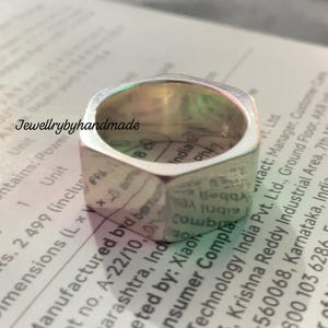 May include: A silver ring with a distinctive hexagonal shape. The ring has a polished, smooth surface and a broad band. The text "Jewellrybyhandmade" is visible in the background. This ring is a piece of jewellery.
