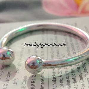 May include: A silver-colored bangle bracelet with a rounded design and two spherical end caps. The bracelet is displayed on a printed surface, with the text "Jewellrybyhandmade" visible. The bracelet is a piece of jewelry.