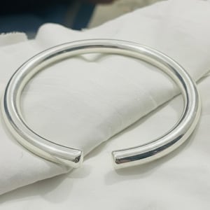 Puede incluir: Un brazalete plateado de extremo abierto. El brazalete tiene un diseño liso y redondeado y un acabado pulido. Los extremos abiertos permiten un fácil uso. El brazalete está sobre una superficie de tela blanca.