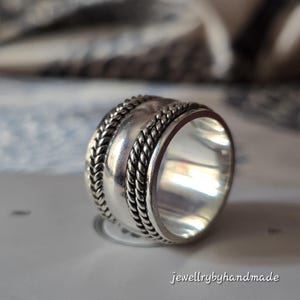 May include: A silver ring with a thick band and a textured design. The ring features a central smooth section flanked by braided rope-like patterns. The ring is displayed on a light-colored surface, with the text "jewellrybyhandmade" visible.