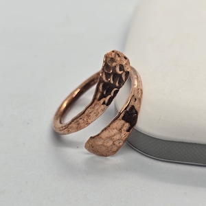 Puede incluir: Un anillo de color cobre con una textura martillada. El anillo se envuelve alrededor del dedo con un diseño abierto. La textura martillada añade un toque rústico y artesanal a la joya.