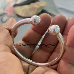 May include: A silver-toned bracelet with two iridescent, spherical accents. The open cuff design features a smooth, polished finish. The text "Jewellrybyhandmade" is visible in the image. This piece is a modern jewelry accessory.